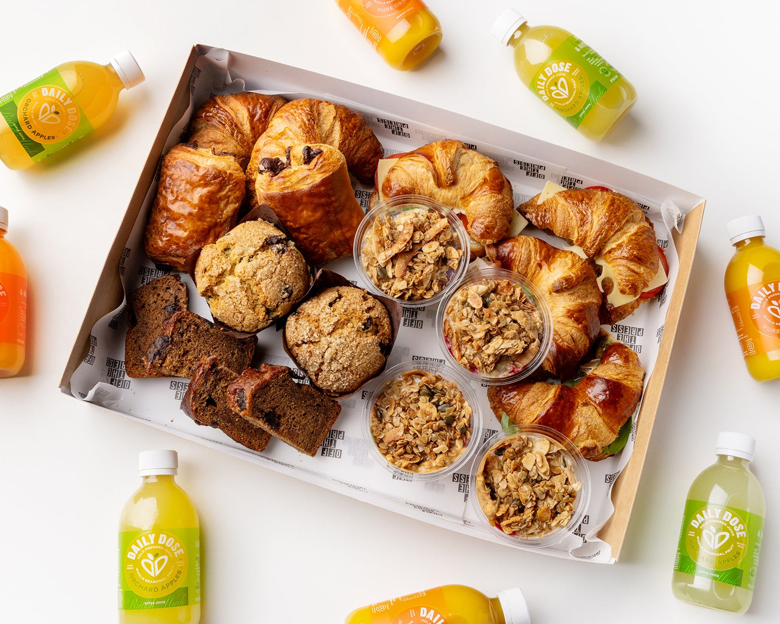 Assorted pastries in a box with small containers of jam and bottles of juice on a white background